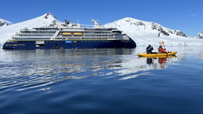 A yellow kayak with two people in front of a blue Lindblad-National Geographic ship and snowy mountains.