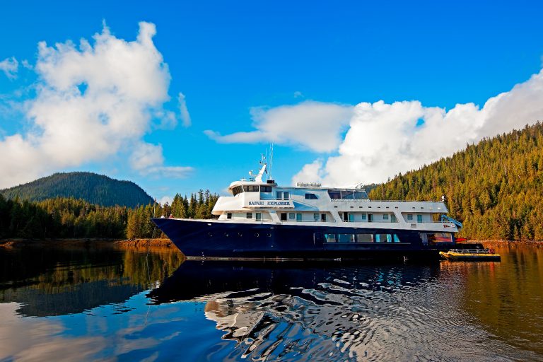 A dark blue and white Safari Explorer ship is on the water, with green, forested mountains in the background and a blue sky with scattered clouds above.
