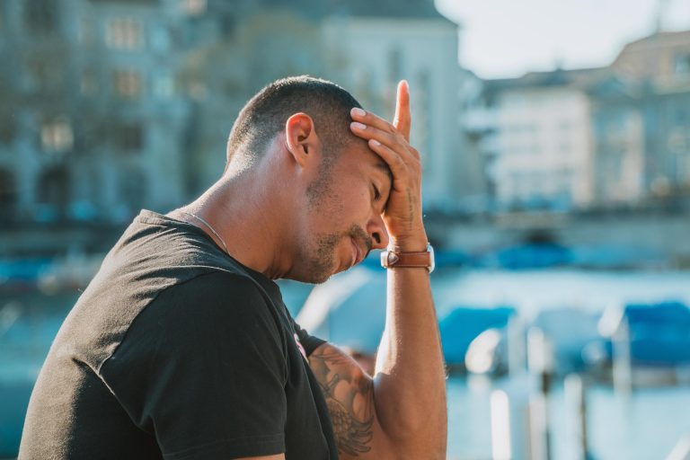 A man with his hand on his forehead, looking stressed or frustrated outdoors in a city setting near water.