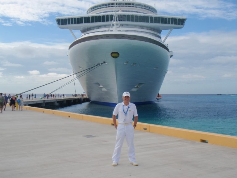 A man in all-white attire stands on a pier in front of a massive, docked cruise ship under a cloudy sky.