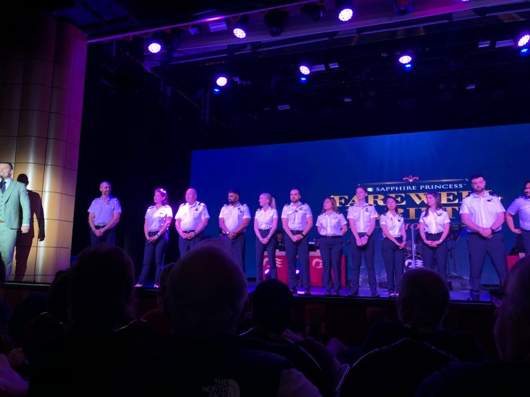 The crew of the Sapphire Princess cruise ship stands on a stage during a farewell show under purple lights.