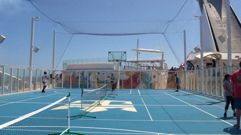 People play pickleball on a blue Royal Caribbean branded court on a cruise ship deck under a netted enclosure.